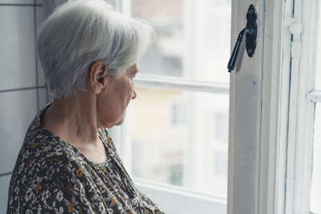 miserable grey-haired older woman looking at the street throughout the window glass. High quality photoの写真素材