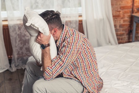 caucasian middle-aged man showing his irritation and screaming out loud but covering his face with a white pillow. High quality photoの写真素材