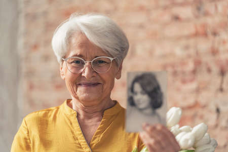 Grinning caucasian white-haired grandma wearing glasses and showing photo of her younger self. High...
