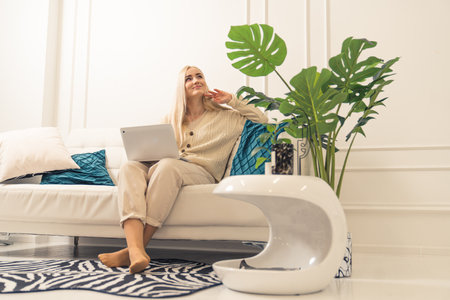 Smiling young woman using laptop, sitting on couch in her modern full of light flat. High quality photoの写真素材