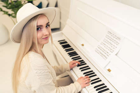 European female blonde long-haired composer in a hat sitting, playing the piano, and looking at camera. High quality photoの写真素材
