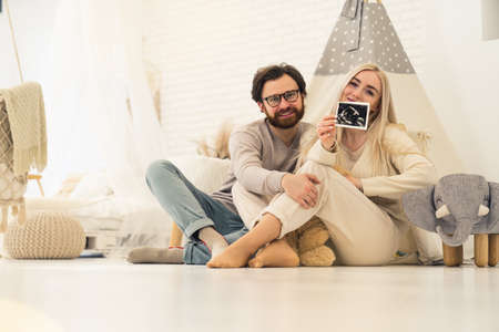 soon-to-be caucasian parents sitting on the floor and showing to the camera an ultrasound scan of pregnancy. High quality photoの写真素材