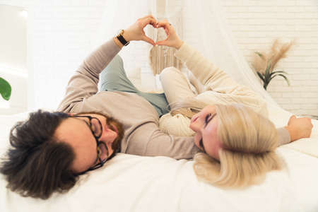 young caucasian couple laying on their bed with white sheets and putting their hands together creating a heart gesture. High quality photoの写真素材