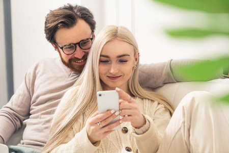 European millennial couple looking cheerfully at a smartphone while sitting together on a sofa. High quality photoの写真素材