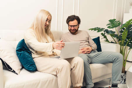 Young caucasian woman with blond hair sitting at home on couch and using laptop. Nearby sits her bearded husband and also looks at the screen. High quality photoの写真素材