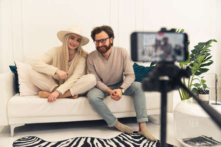 young caucasian influencer couple vlogging with a smartphone while sitting on a white sofa in their modern apartment. High quality photoの写真素材
