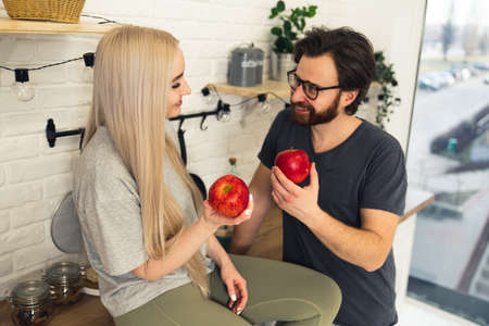 cute couple in their apartment sharing good habits and eating one apple a day, promoting healthy eating while sitting in their modern kitchen. High quality photoの写真素材