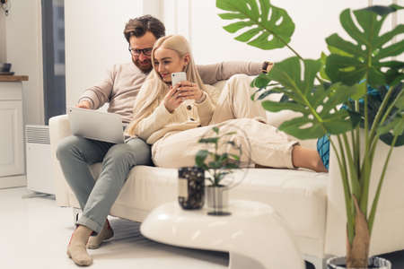 cute young adults couple with laptop and smartphone sitting on white couch and enjoying networking and social media. High quality photoの写真素材
