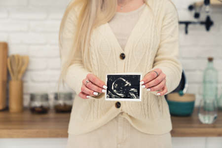 single caucasian mother showing ultrasound picture of her baby to the camera while standing in he kitchen. High quality photoの写真素材
