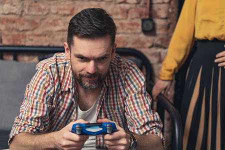 caucasian man in an unbuttoned checked shirt playing games with blue game controller and ignoring his woman friend. High quality photoの写真素材