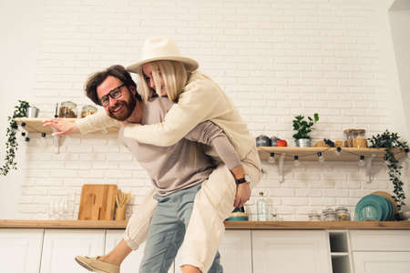 caucasian middle-aged bearded man having fun and lifting his fiancee piggyback in their modern bright kitchen. High quality photoの写真素材