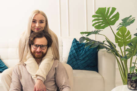 young beautiful caucasian long-haired blonde woman hugging her middle-aged husband with facial hair from the back, looking at camera. High quality photoの写真素材