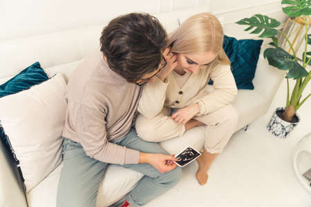 Young caucasian couple looking at their baby ultrasound picture while sitting on a white sofa in their modern apartment. High quality photoの写真素材