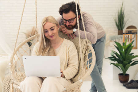 young millennial couple watching videos together on a white laptop while sitting on a modern swing. High quality photoの写真素材