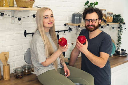 young married caucasian couple sharing healthy lifestyle, holding a red apple while being in the kitchen and looking at camera. High quality photoの写真素材