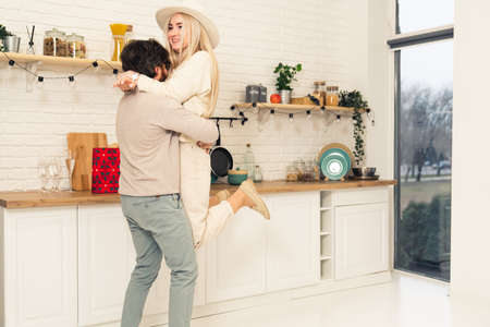 millennial caucasian brunet man lifts up his beautiful blonde long-haired wife in their modern kitchen while celebrating. High quality photoの写真素材