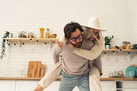 European spouses in their bright modern kitchen having fun and laughing, man lifting his female partner on his piggyback. High quality photoの写真素材