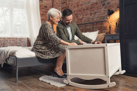 European elderly grandmother and her millennial son swing the white crib where a little infant baby is laying. High quality photoの写真素材