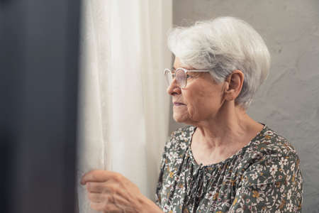 portrait upset caucasian older woman pensioner lady touching curtain gazing through the window. High quality photoの写真素材