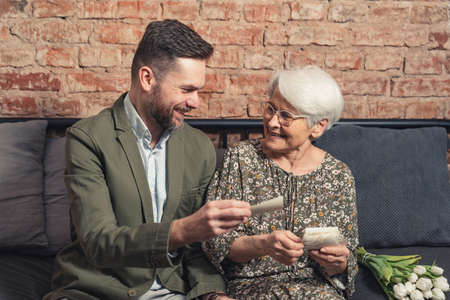 Portrait of laughing grey-haired grandmother and smiling young handsome man looking at photos in his flat. High quality photoの写真素材