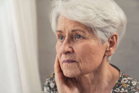 Old caucasian sad lady looking through the window with a melancholic gaze. High quality photoの写真素材