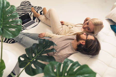 two caucasian people in a relationship laying down, looking at each other, holding laptop between their legs. High quality photoの写真素材