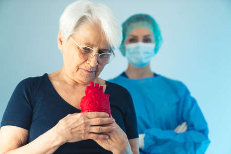 Elderly person standing in front of a surgeon, holding artificial red heart in front of her chest with sad worried expression. High quality photoの写真素材