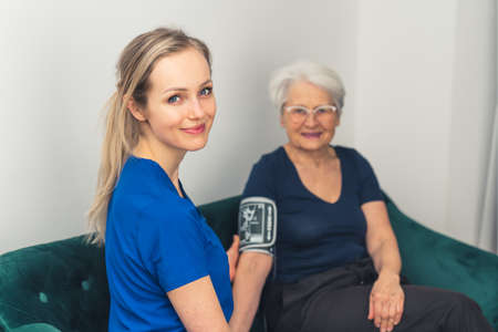 Friendly caregiver and her elderly patient sitting together on a sofa, measuring pressure, and looking at camera. High quality photoの写真素材