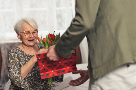 pleasantly surprised elderly grey-haired pensioner woman receiving a bouquet and a gift from her...
