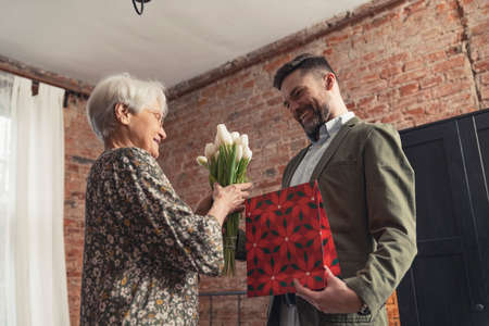 adult handsome son with facial hair giving red gift and white tulip bouquet to his elderly mother. High quality photoの写真素材