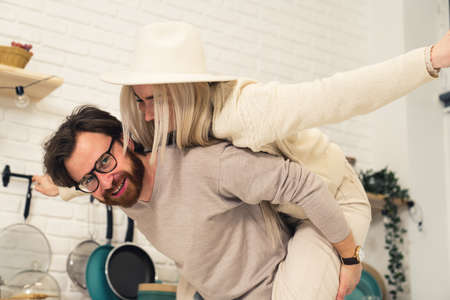 Portrait of young Caucasian couple woman sitting on husbands back man smiling to the camera kitchen background. High quality photoの写真素材