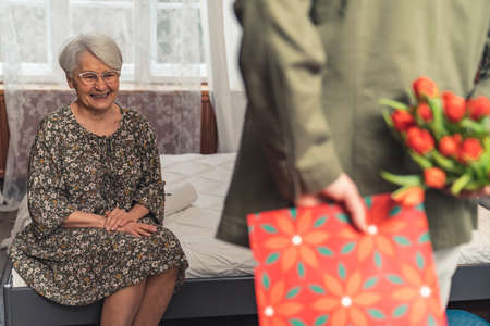 back view grandson hiding presents for his grinning grey-haired caucasian pensioner grandmother ....