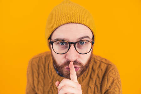 Closeup of young bearded Caucasian man looking at the camera showing hush shh gesture isolated orange background focus on the head . High quality photoの写真素材