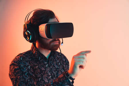 caucasian man using googgles for virtual reality and pointing in a direction in front of him. Medium Close up shot. Orange Background. High quality photoの写真素材