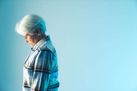 resigned elderly gray-haired grandmother lowering her head in disappointment, side view medium studio shot with blue background. High quality photoの写真素材