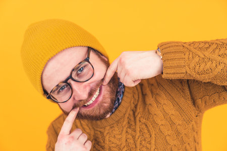 Portrait of smiling young Caucasian man in glasses, hat and beard, man touching his edges of mouth, showing teeth orange background isolated studio shot . High quality photoの写真素材