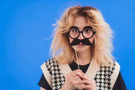 Playful goofy teenage caucasian girl holding fake black mustache near her face, isolated on blue background. High quality photoの写真素材