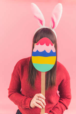 Vertical studio shot caucasian woman covering her face with colorful cardboard Easter egg while...