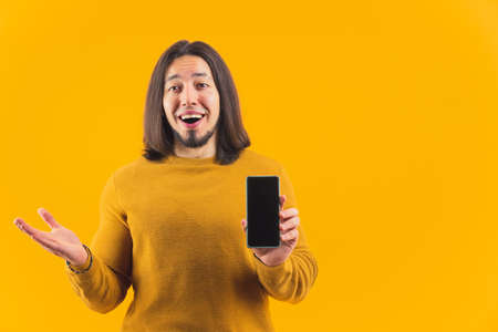 Caucasian bearded man surprised and amazed by huge sales holding mock-up black screen smartphone. Studio shot over yellow background. High quality photoの写真素材