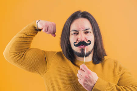 Goofy muscled caucasian man in his 30s doing silly faces and covering his face with fake moustache while looking at camera. High quality photoの写真素材