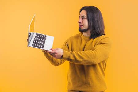 Disgusted angry caucasian guy with facial hair holding laptop up and looking at the screen. Studio shot over yellow background. High quality photoの写真素材