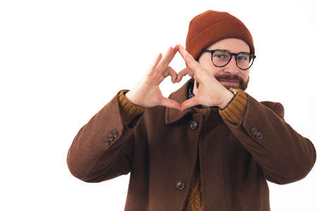 Portrait of young bearded Caucasian man in glasses and hat smiling and showing heart gesture with...