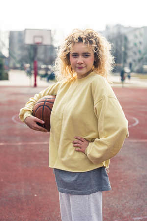 Confident young blond Caucasian woman stands in a light yellow sweater on the sports field holding a basketball ball and keeping her another hand on her waist medium full shot . High quality photoの写真素材