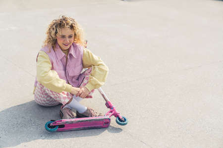 Pretty young blond curly haired hipster woman sits on the ground smiling, legs crossed, pink scooter in front of her looking away full shot copy space gray background . High quality photoの写真素材