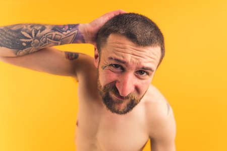 Studio portrait of a bare chested caucasian guy covered with various tattoos looking confused at camera and scratching his head over yellow background. High quality photoの写真素材