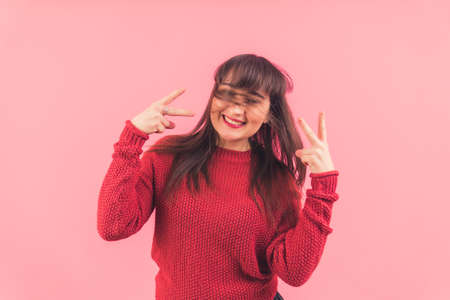Happy funny European brunette with hair covering her face showing v-sign to the camera and smiling. Pink background, studio shot. High quality photoの写真素材
