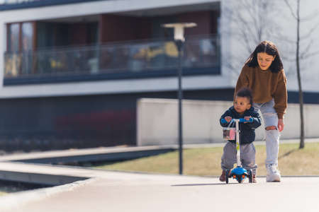 black-haired thin woman following a little African-American boy with curly hair standing on the scooter and looking sideways full shot urban background. High quality photoの写真素材