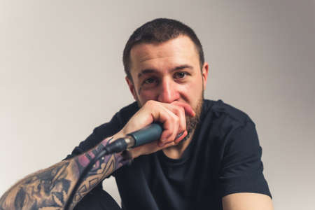 attractive brown-eyed man with a tattoo sitting holding a microphone in his hand and putting on mouth medium closeup light background studio shot. High quality photoの写真素材