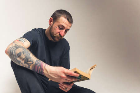 handsome bearded Caucasian tattooed man sitting on the floor and reading the Holy Bible - faith concept isolated on light background medium full shot studio shot. High quality photoの写真素材
