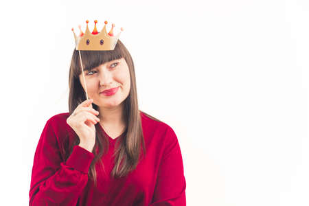 Content pensive caucasian young funny woman holding fake paper crown above her head. Medium studio shot over white background. High quality photoの写真素材
